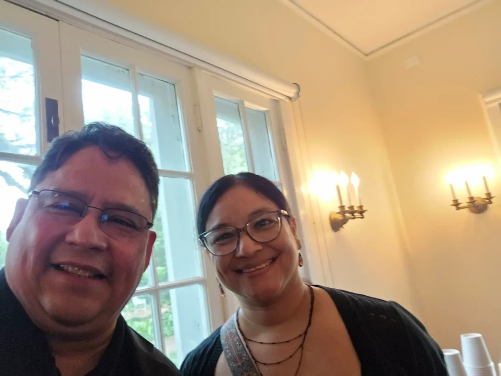 Two people smiling indoors near large windows with white frames. Warm light from wall sconces illuminates the room. Both individuals are wearing glasses and dark clothing, and greenery is visible outside the windows.