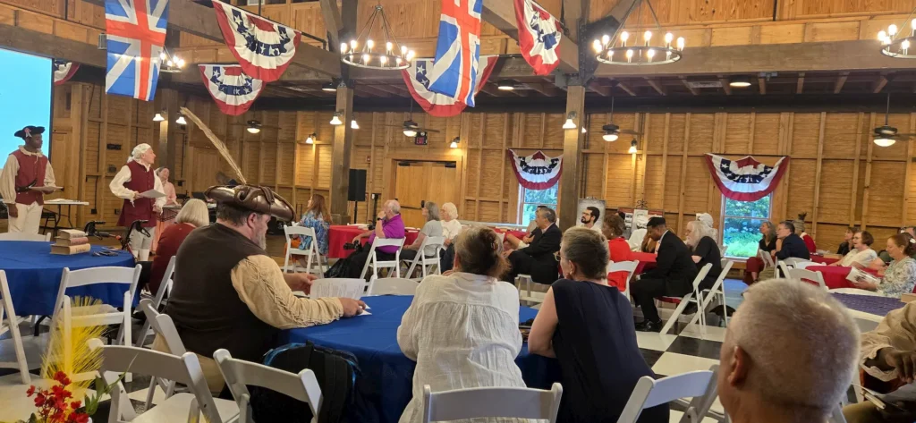 People attend an indoor event with colonial-era reenactors speaking at the front. The hall is decorated with patriotic bunting and flags, and attendees sit at tables covered in blue and red tablecloths.