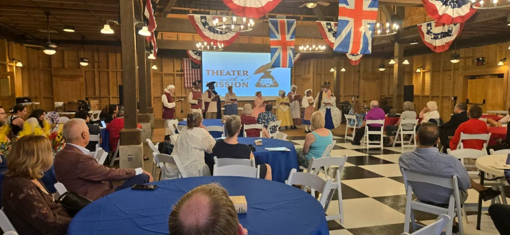 People sit at round tables watching a stage performance in a large, wood-paneled hall decorated with patriotic bunting and large British flags. Performers on stage wear historical costumes, and a screen reads "THEATER with a MISSION.