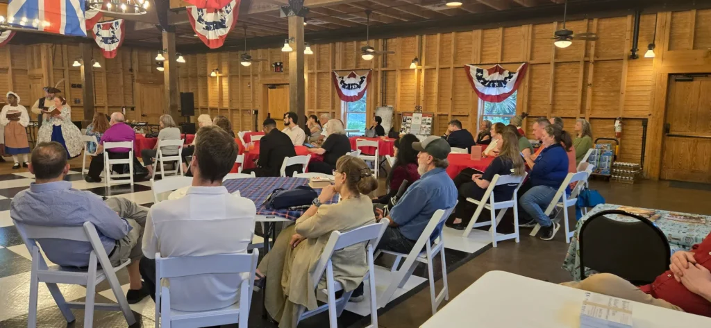 A group of people sit on white chairs around tables with red tablecloths in a wooden hall decorated with patriotic banners, listening to a speaker at the front.