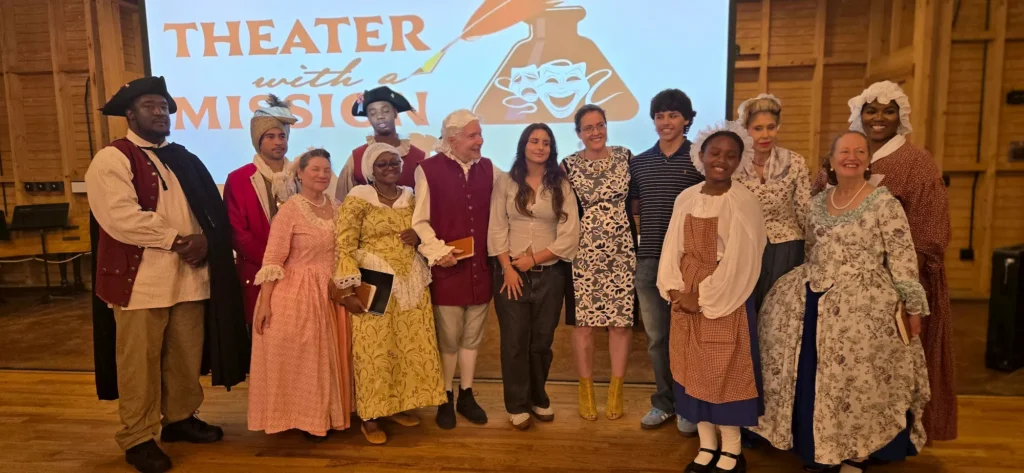 A diverse group of people in historical costumes and modern clothing pose smiling on a wooden stage in front of a screen that reads "THEATER with a MISSION" with theater mask graphics.