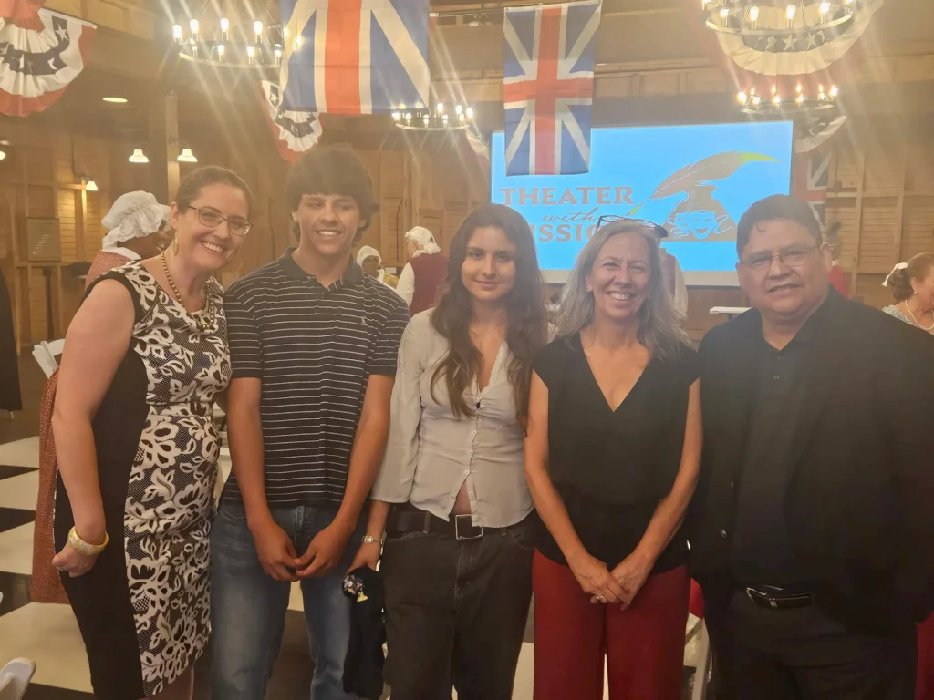 Five people are standing and smiling indoors at an event with British flags and classic decorations. A screen in the background reads "THEATER" with a comedy and tragedy mask logo.
