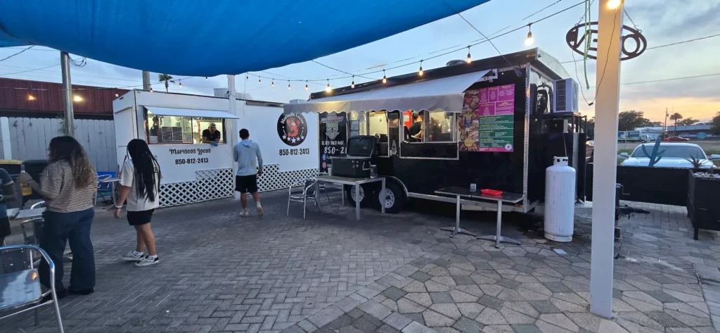 A food truck and a small white order booth are set up on a paved patio at dusk. Several people stand nearby, and string lights hang overhead, creating a casual outdoor dining atmosphere.