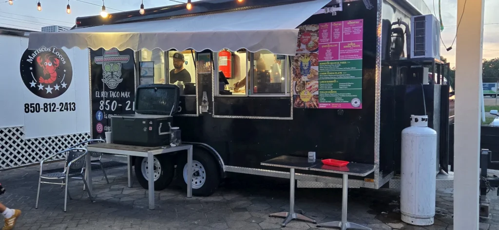 A black food truck with colorful menus and signage is parked outdoors at dusk. Two workers are visible inside. A table with a cooler and chairs is set up in front, with string lights overhead and a propane tank nearby.