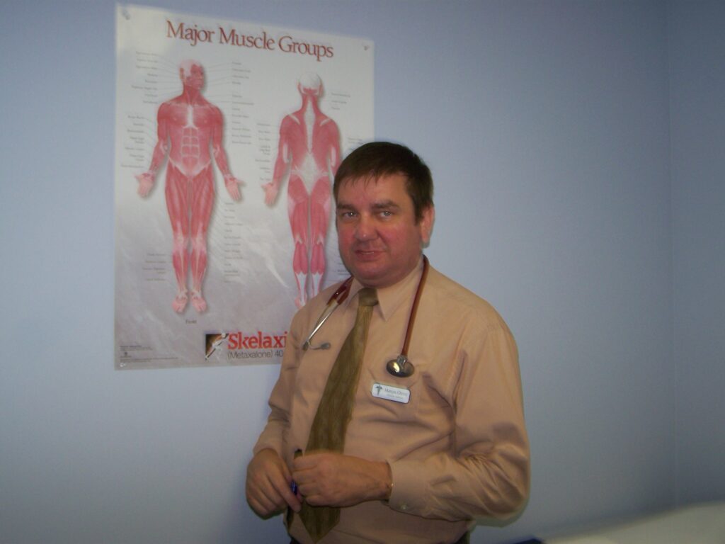 PA. Marcos Otero, with a stethoscope around his neck, stands in front of a poster showing major muscle groups in a medical examination room.