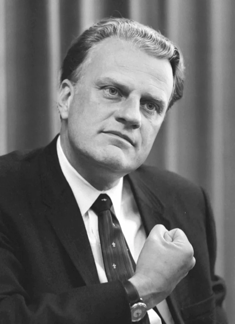 Black and white photo of a man in a suit and tie, looking thoughtful and resolute, raising his clenched fist slightly in front of him, with curtains in the background.