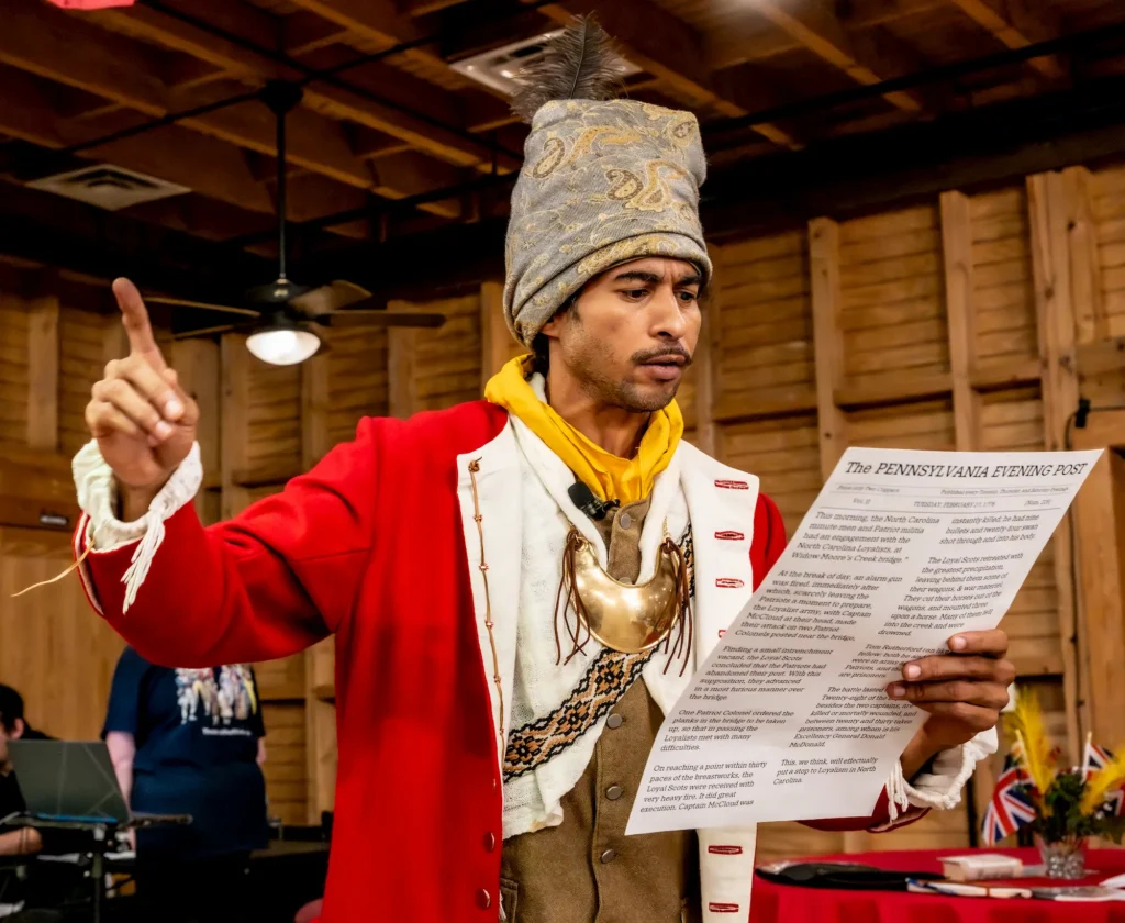 A man in historical attire, including a tall embroidered hat and red coat, gestures with one hand while reading from a paper titled "The Pennsylvania Evening Post" inside a wooden room.