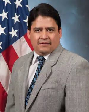 David Triana, wearing a light gray suit, white shirt, and patterned tie, stands in front of a U.S. flag and a gray-blue background, looking at the camera.
