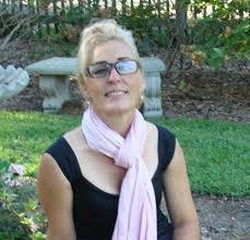 Deborah Desilets, a woman with blonde hair and glasses, sits outdoors on grass wearing a black top and light pink scarf. A stone bench and lush greenery provide a serene backdrop.