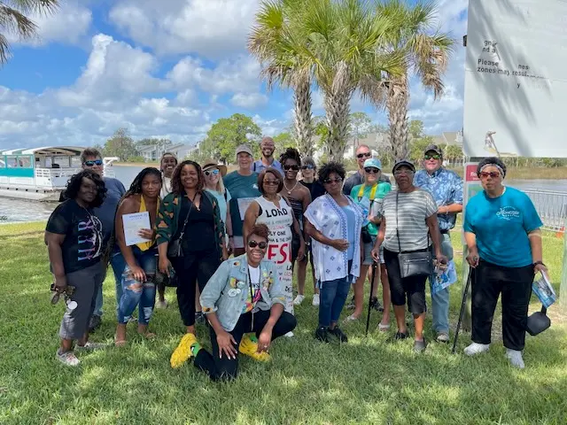 A diverse group of people poses together outdoors on green grass, with palm trees and blue sky in the background. Some hold water bottles and papers, appearing cheerful and relaxed on a sunny day.