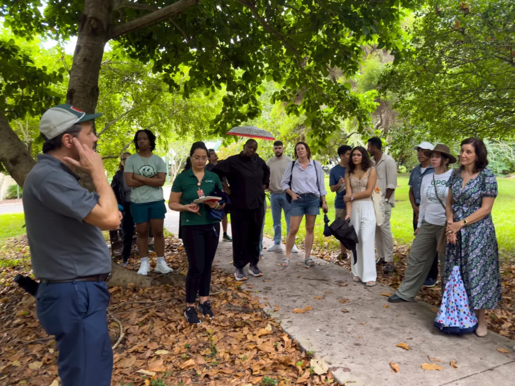 A group of people stands and listens to a guide speaking under a large tree on a leafy path. Some people hold umbrellas or notebooks, and most are casually dressed, gathered in a shaded outdoor setting.