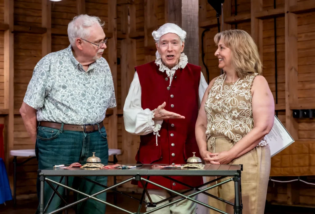 Three adults stand indoors by a table with bells and ribbons. The man in the center wears historical colonial attire, while the other man and woman wear modern casual clothes. They appear to be in conversation and smiling.