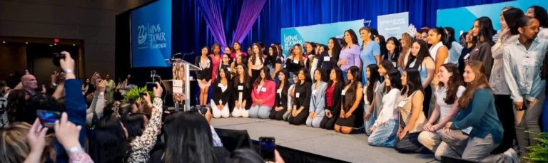 A large group of women and girls pose together on stage at an event, while people in the audience take photos. The stage features blue curtains, banners, and a podium.