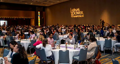 Un gran grupo de personas sentadas en mesas redondas en una sala de conferencias durante el Simposio Latinas Power, con el logotipo del evento proyectado en la pared.