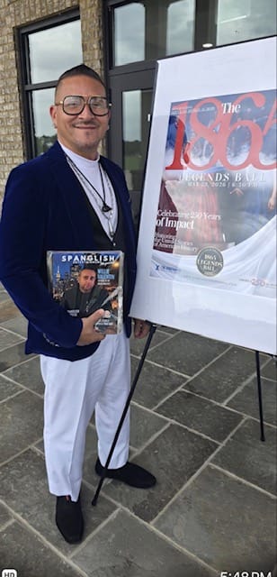 A smiling person in a blue blazer and white pants stands next to a poster on an easel and holds a “Spanglish” magazine with their own photo on the cover. They are outside a building with large glass doors.