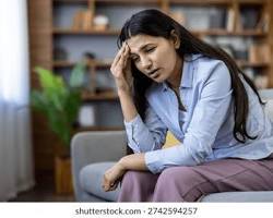A woman sits on a couch with her hand on her forehead, appearing stressed or in pain from a headache, in a living room with a blurred background of shelves and a plant.