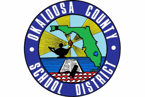 The seal of Okaloosa County School District featuring a map of Florida, a lit oil lamp, an open book, and rays of light, all within a blue circular border with the district name.