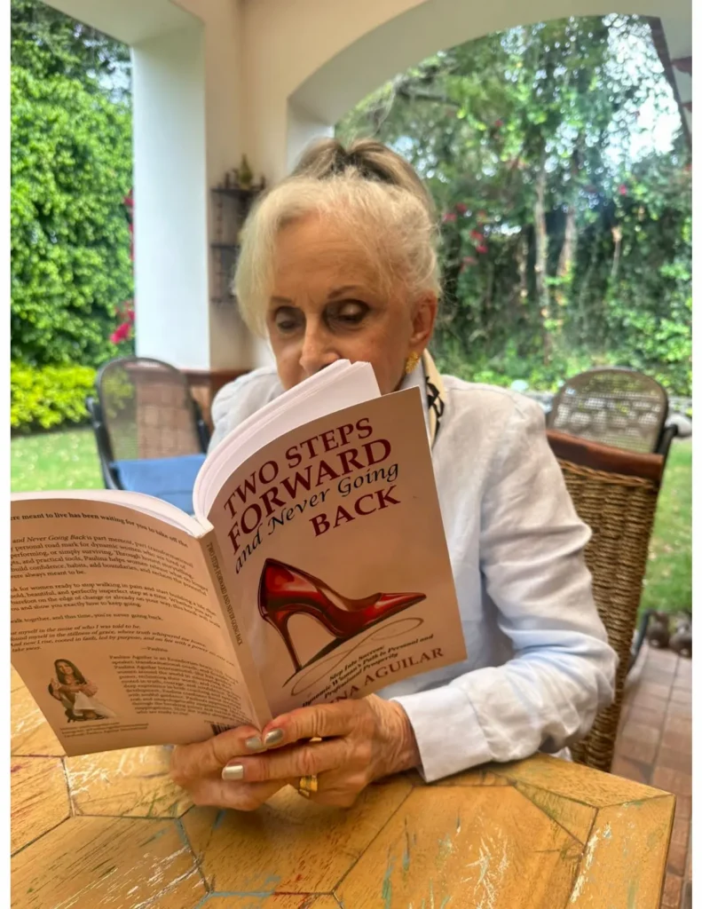 An elderly woman with white hair sits outdoors at a table, reading a book titled "Two Steps Forward and Never Going Back" with a red high heel on the cover. Lush greenery and patio furniture are in the background.