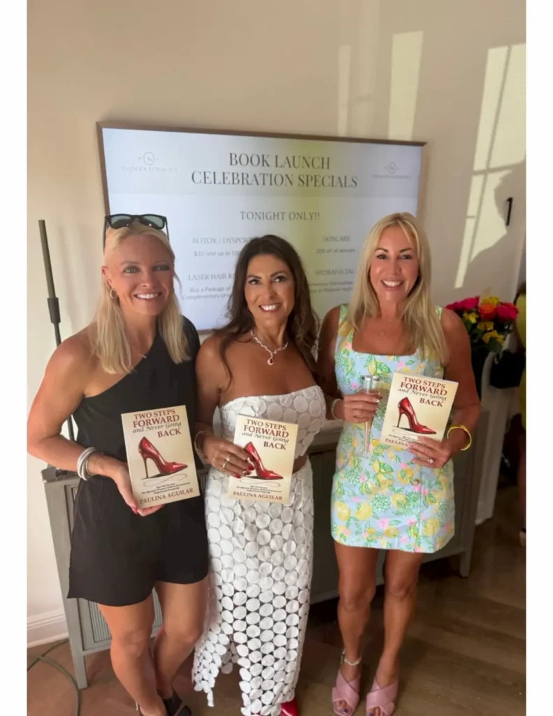 Three women standing indoors, smiling and holding copies of a book titled "Forward, Back," with a high-heeled shoe on the cover. A book launch event sign and a bouquet of flowers are visible in the background.