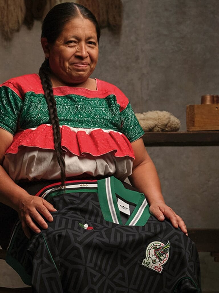 An older woman with braided hair wears a red, green, and white traditional blouse and smiles while sitting indoors, holding a black Mexico soccer jersey with the team's logo.