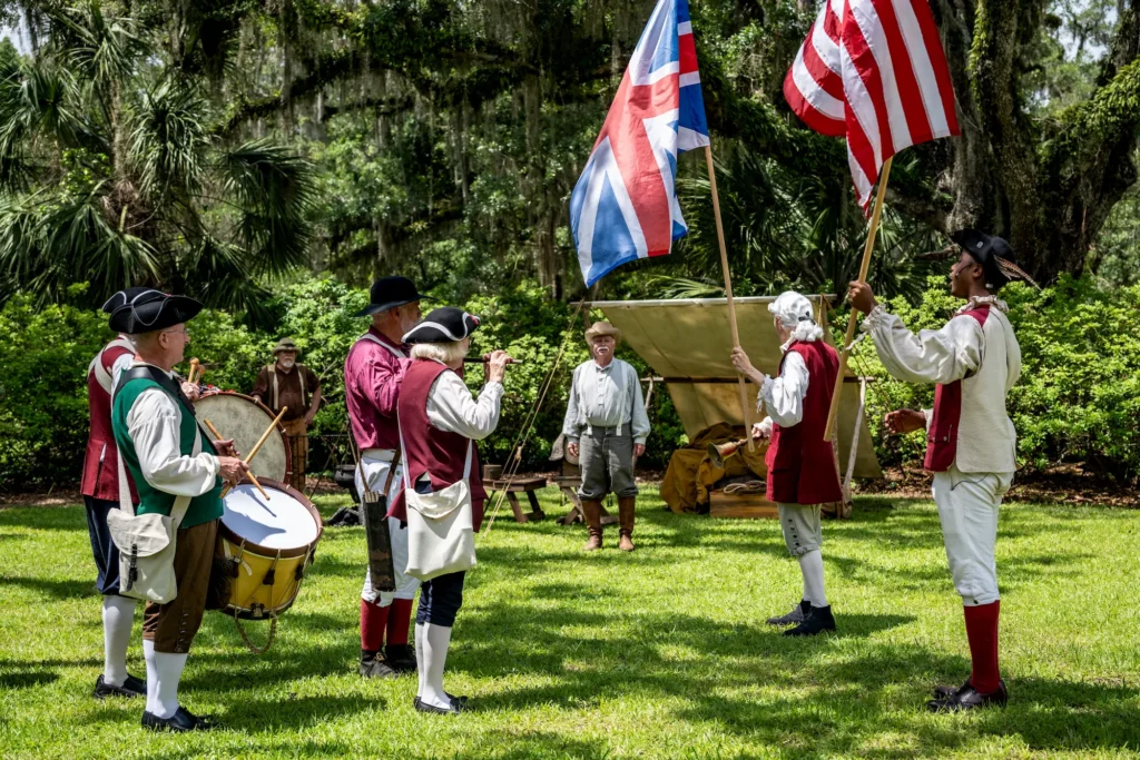 A group of people in colonial-era costumes reenact a historical scene outdoors. Two hold flags, one holds a drum, and others stand nearby. Trees and a tent are visible in the background on a sunny day.