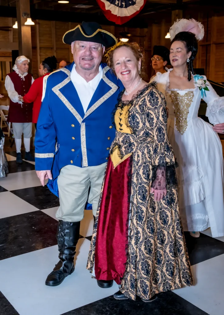 A smiling man and woman pose in historical costumes at an indoor event; he wears a blue colonial military uniform with a tricorne hat, and she wears a gold and black patterned gown. Other costumed people stand in the background.