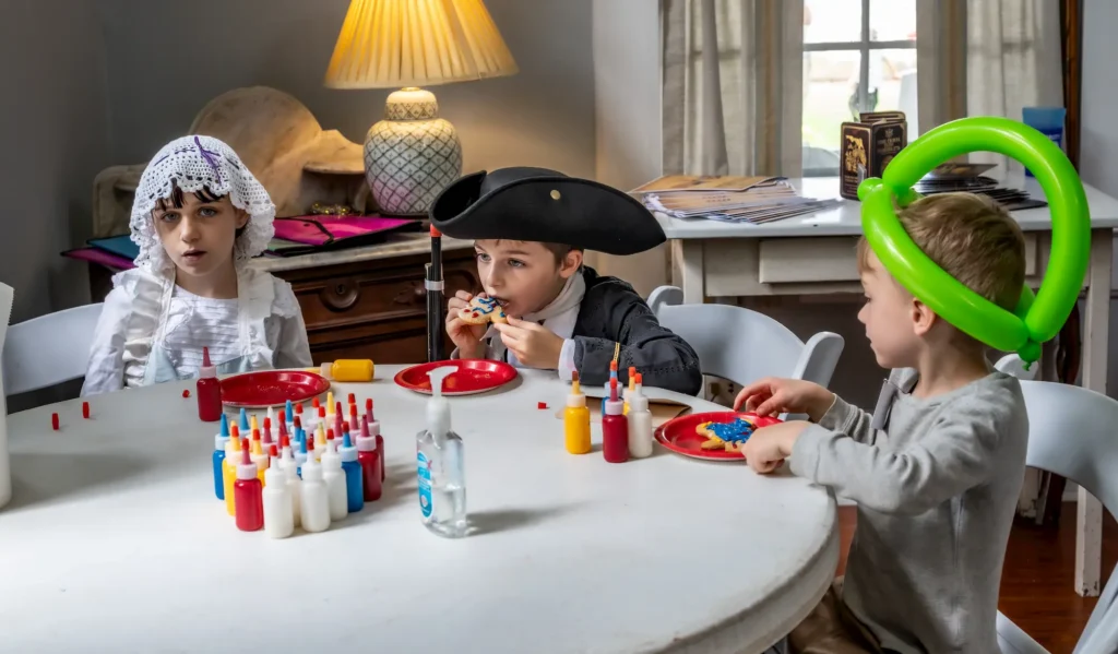 Three children in costumes sit around a table decorated with bottles of paint and cookies. One eats a cookie, another wears a lace bonnet, and the third wears a green balloon hat. There is a lamp and papers in the background.