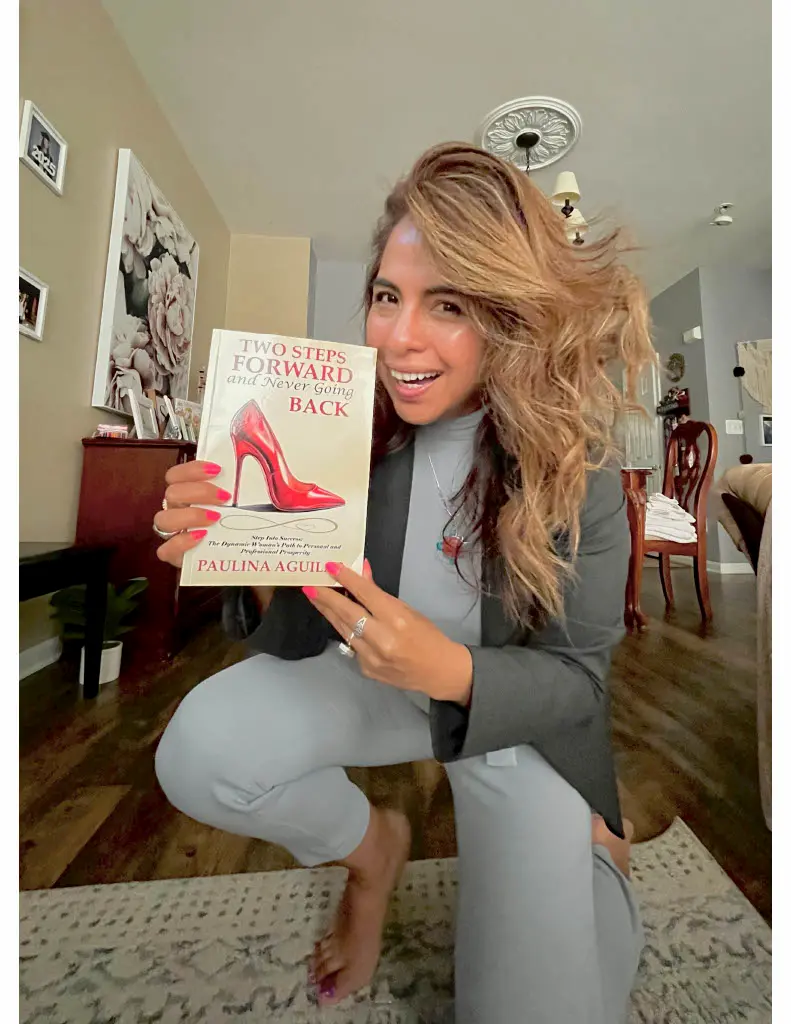 A smiling woman with long, wavy hair and a gray outfit kneels indoors, holding up a book titled "Two Steps Forward and Never Going Back" by Paulina Aguilar. The background shows a living room with decor and furniture.