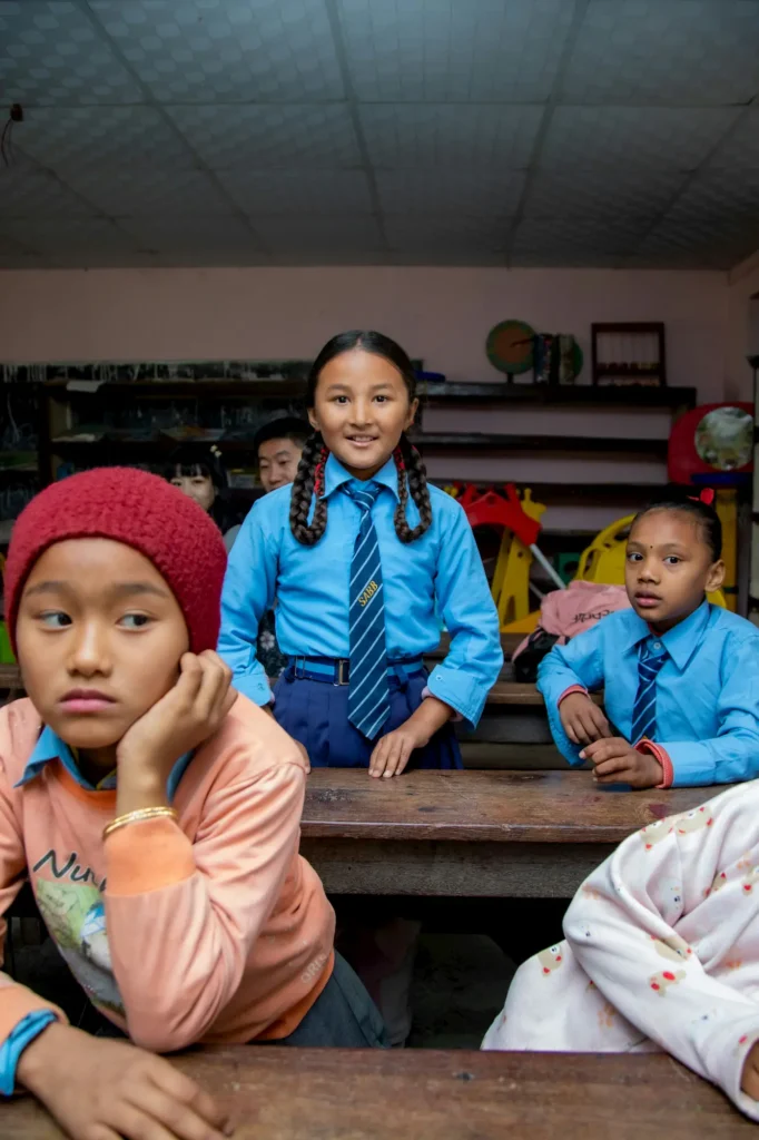 A smiling girl in a blue school uniform stands between desks in a classroom, with other children seated around her. One boy in a red beanie rests his face on his hand, while another looks to the side.