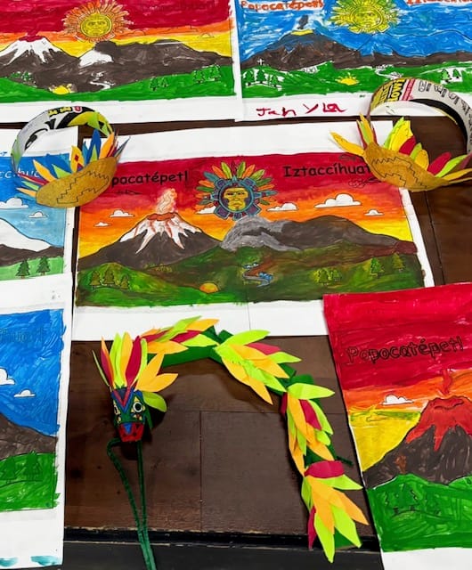 Colorful handmade drawings of mountains, crowns, and a feathered serpent mask are displayed on a table at Un Poquito de Mexico Summer Camp in Quincy. The artwork features bright reds, blues, and greens, and some pieces are labeled “Popocatépetl” and “Iztaccíhuatl.”.