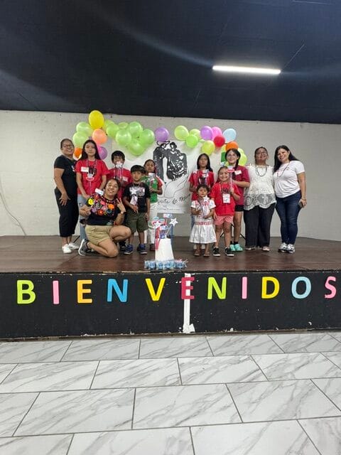 A group of children and adults pose on a stage decorated with balloons and a welcome sign that says "BIENVENIDOS" at the Mexico Summer Camp in Quincy. The children hold up small prizes, and everyone is smiling, celebrating their time together at Un Poquito de Mexico.