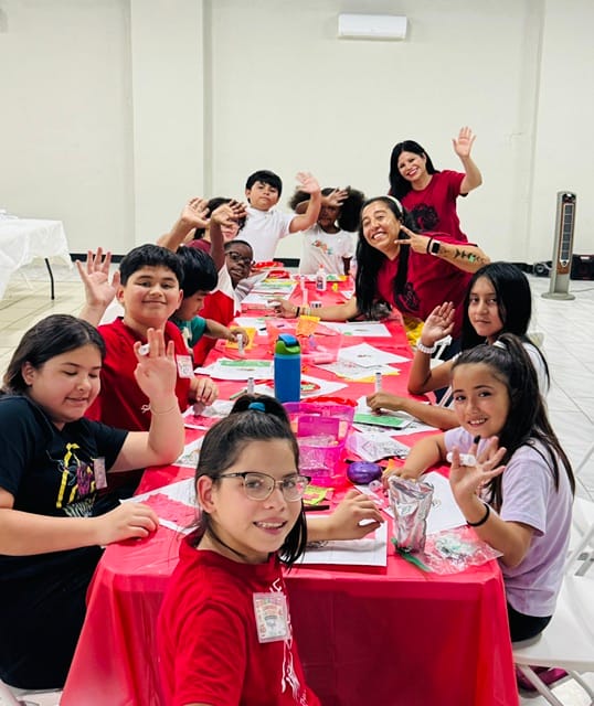 A group of smiling children and one adult sit around a red-covered table, waving at the camera. The table is filled with art supplies and snacks, and everyone appears to be enjoying a creative activity indoors at Summer Camp Quincy, inspired by Un Poquito de Mexico.