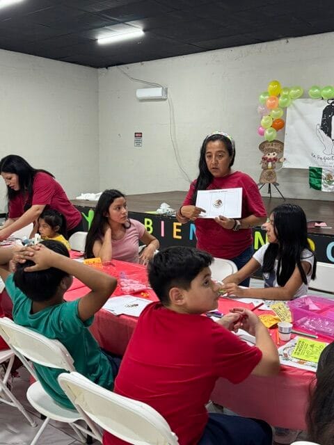 A woman holds up a booklet while speaking to a group of children and adults seated at a table decorated with craft supplies during an Un Poquito de Mexico Summer Camp in Quincy. Balloons and a bear-themed banner are visible in the background.