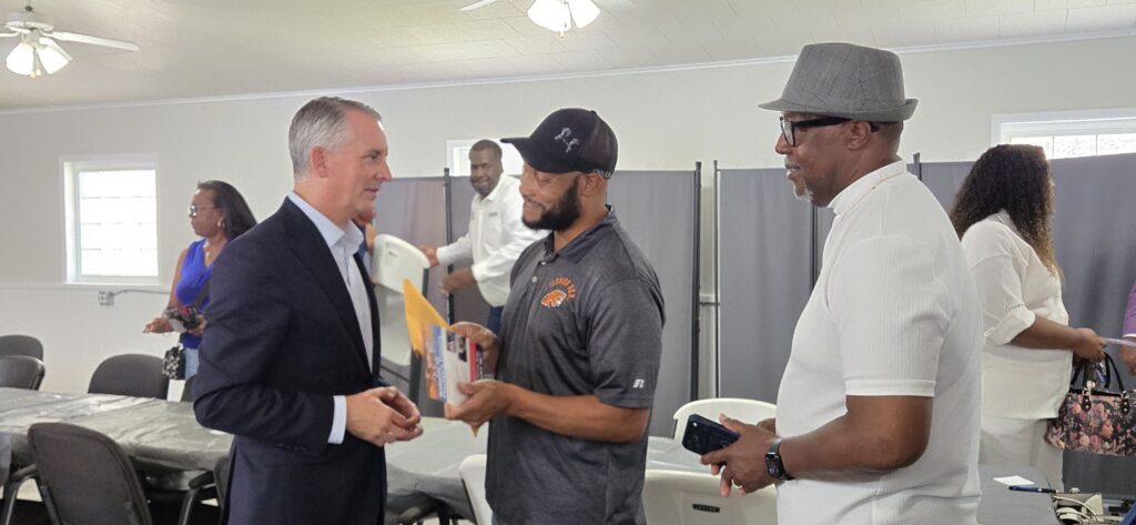 A man in a suit shakes hands with another man holding papers, while a third man in a hat stands nearby. Several other people are in the background of the room.
