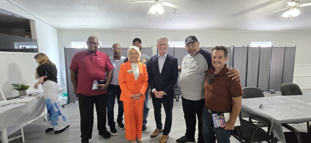 A group of seven people pose together in a room with white walls and tables; one woman in an orange suit stands in the front center, surrounded by six men.