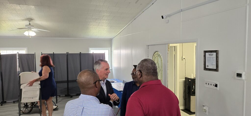 Four men stand talking inside a room with white walls, while a woman in a blue dress walks near privacy screens and a baby stroller. A certificate and a thermostat are visible on the wall.