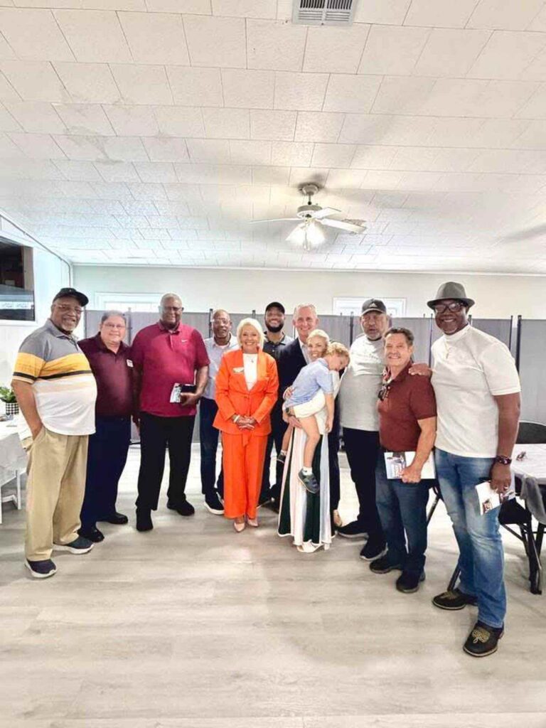 A group of twelve adults and one child stand together indoors, smiling at the camera. One woman in an orange suit stands in the center. The setting appears to be a casual gathering or community event.