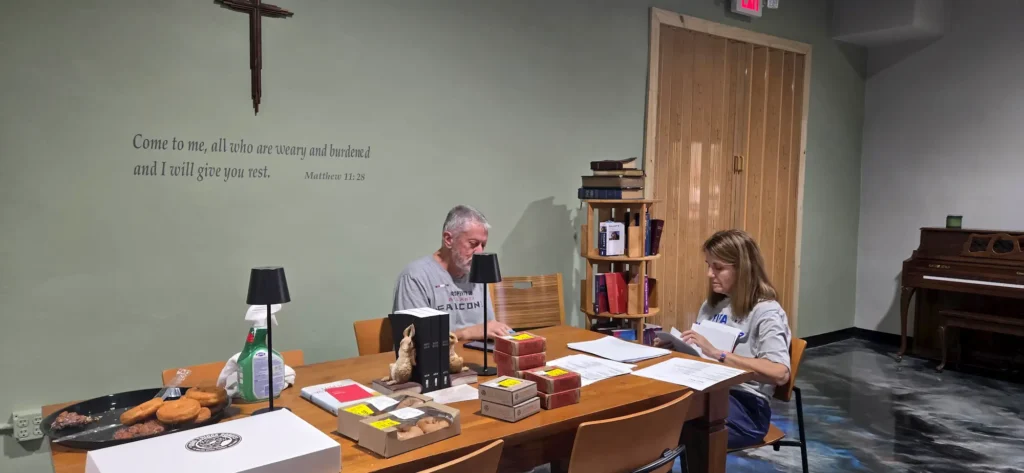 Two people sit at a wooden table with papers, boxes of donuts, and a drink. A Bible verse is on the wall behind them. A cross, piano, and books are also visible in the room.