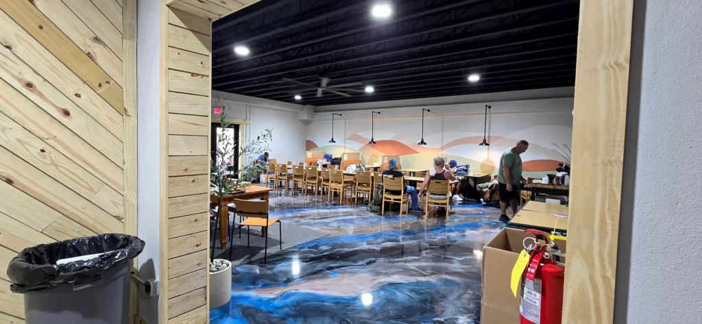 A modern workspace with wooden tables and chairs, people working, a colorful mural on the wall, polished blue and black floor, ceiling lights, and a fire extinguisher near the entrance.
