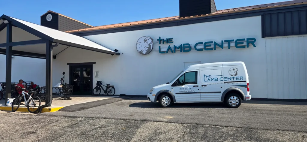 A white van labeled "The Lamb Center" is parked outside a building with the same name and a lamb logo. Several bicycles and people are near the entrance under a shaded awning.