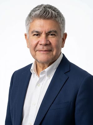 A middle-aged man with short gray hair wearing a navy blue blazer and white shirt, smiling slightly against a plain white background, exuding the warmth and confidence of someone who champions financial dignity—just like Louis Barajas in his work promoting Finances with Heart.
