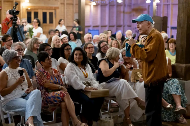 A person in a brown jacket and blue hat speaks into a microphone to a seated audience in a large indoor space during the 10th anniversary of the Micro Theater Festival by Tallahassee Hispanic Theater. The crowd listens attentively, some smiling and others holding items.