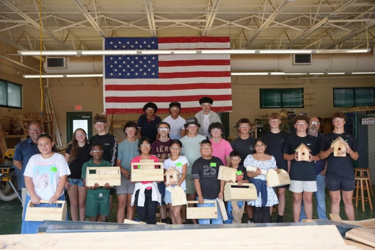 A group of teens wearing safety glasses stand in a workshop holding wooden projects, such as birdhouses and toolboxes, with a large American flag hanging on the wall behind them.