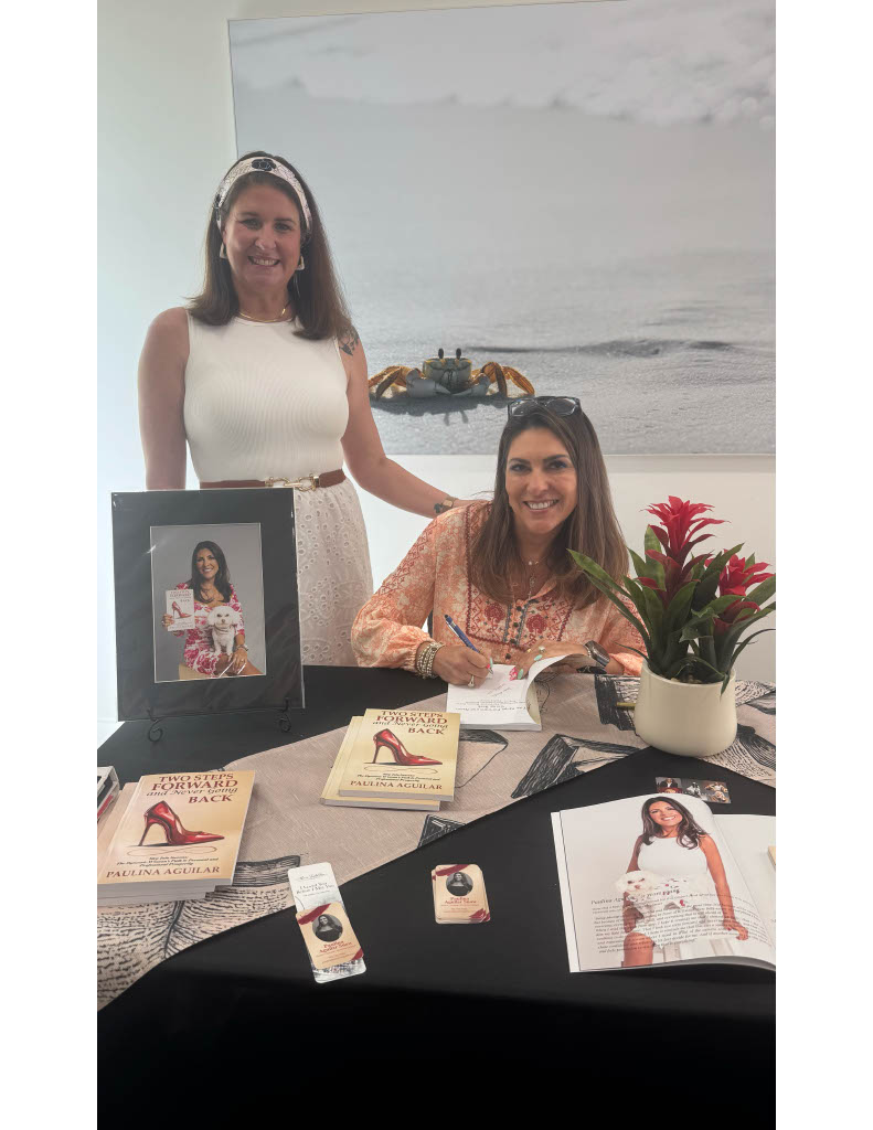 Two women at a book signing table embody authenticity; one is seated, living her truth as she signs a book, while the other stands supportively behind her. Several books, a framed photo, and a plant are arranged on the table. A large beach photo—symbolizing freedom—hangs on the wall behind them.