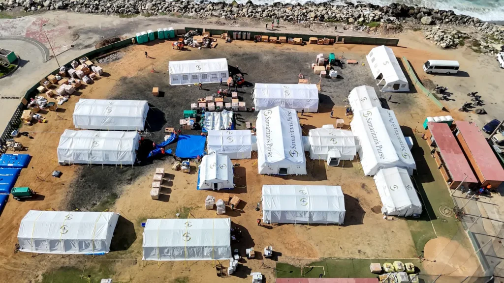 Aerial view of a disaster relief camp with white tents, scattered supplies, and people working. The camp is set up near a rocky shoreline with portable toilets and equipment visible.