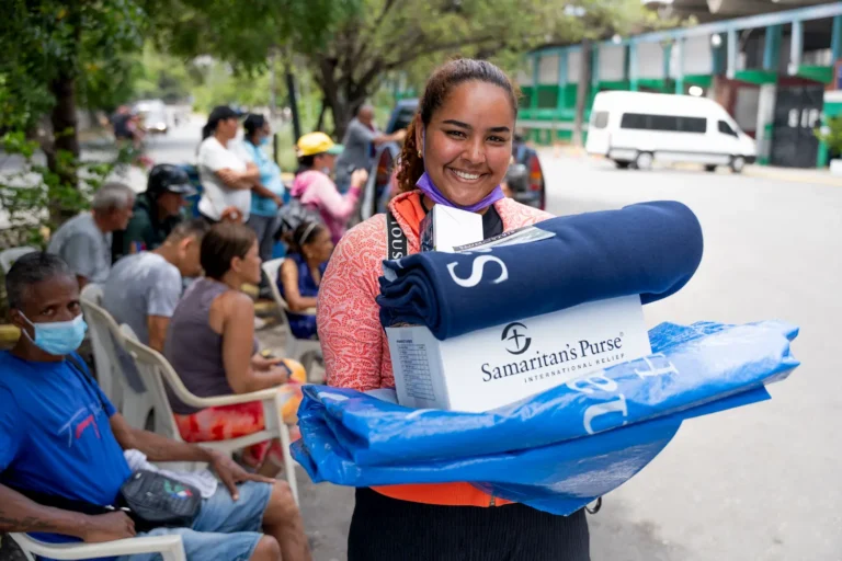 A smiling woman holds a Samaritan's Purse box, a navy blue blanket, and a blue bag outside, with seated people and a white van in the background.