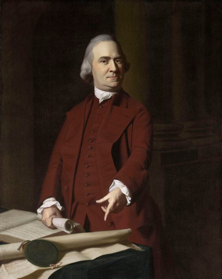 A man in 18th-century attire stands beside a table with papers and a scroll, gesturing with one hand. He has white hair and wears a maroon coat, white shirt, and waistcoat. The background is dark and formal.