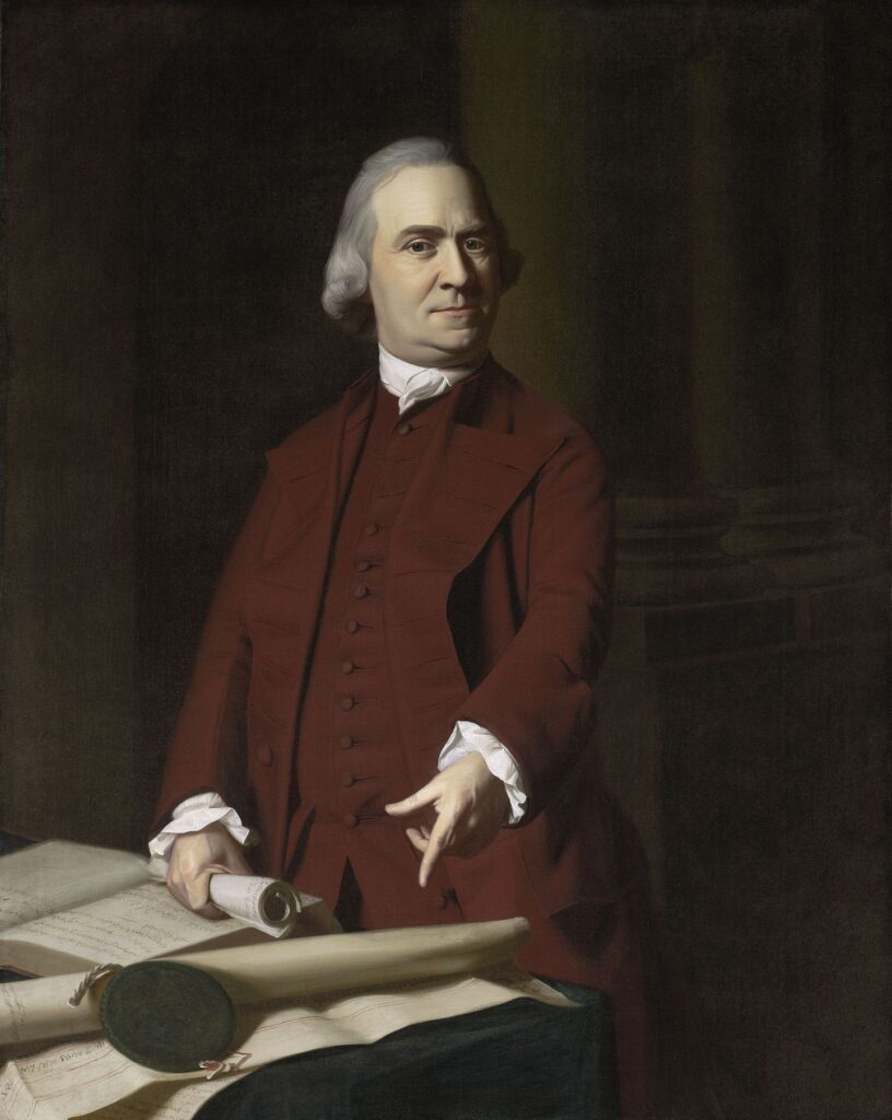 A man in 18th-century attire stands beside a table with papers and a scroll, gesturing with one hand. He has white hair and wears a maroon coat, white shirt, and waistcoat. The background is dark and formal.