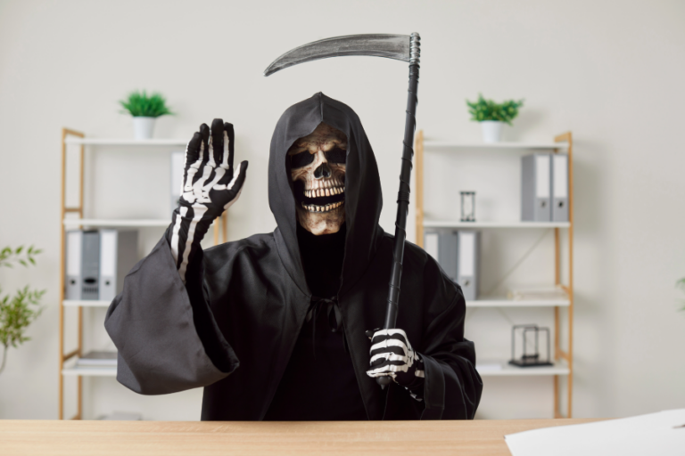 A person dressed as the Grim Reaper, wearing a skull mask and black hooded robe, holds a scythe and waves while sitting at an office desk with shelves and plants in the background.