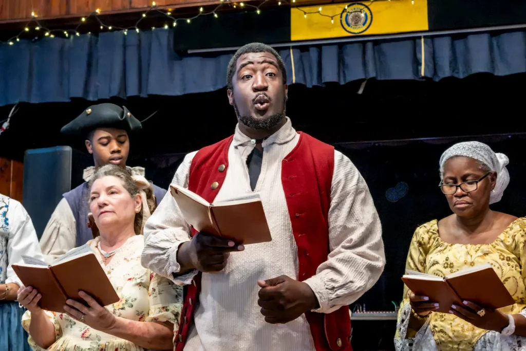 Four people in historical costumes sing from books on stage. One man in a red vest stands centered, singing passionately. Others surround him, also holding books. String lights and a blue curtain hang in the background.